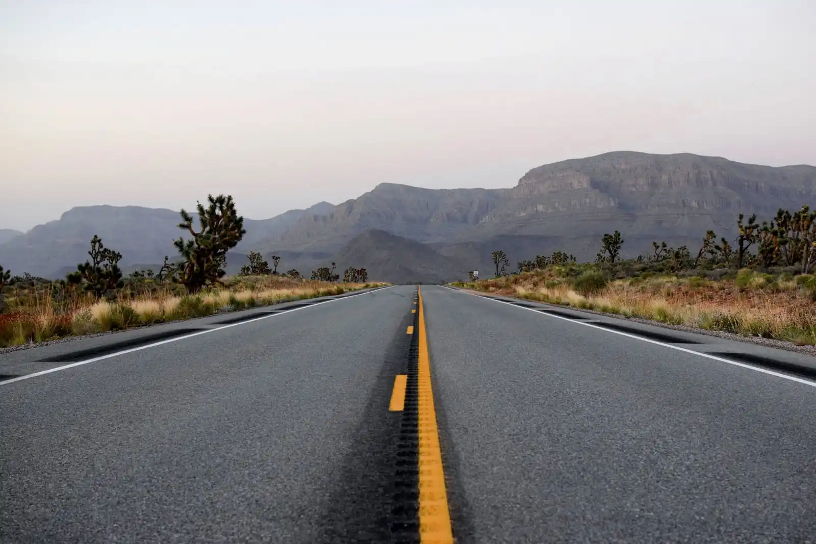 An empty two-lane desert road at dusk running straight toward a layered mountain range
