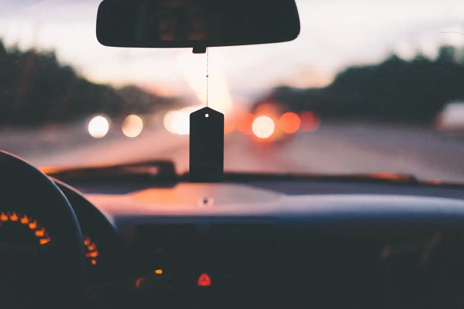 The view over a car dashboard at dusk, the instrument cluster lit amber behind the wheel and a warning lamp burning below it