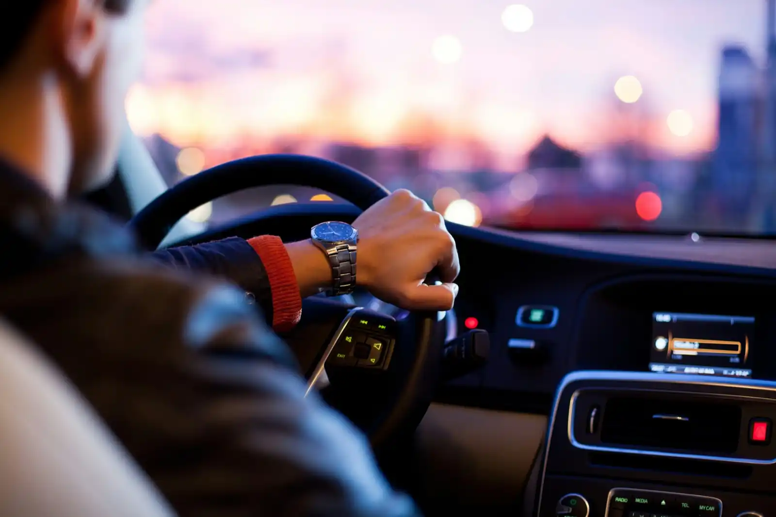A driver’s hand on the steering wheel at dusk with city lights blurred ahead