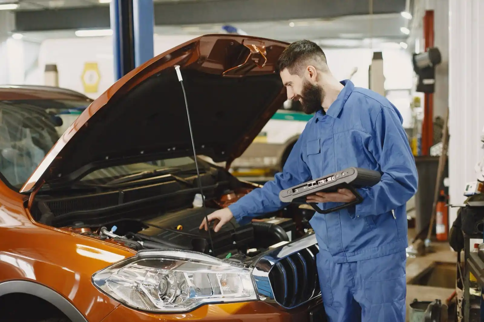 A smiling technician using a diagnostic tablet beside an open hood in an auto repair shop