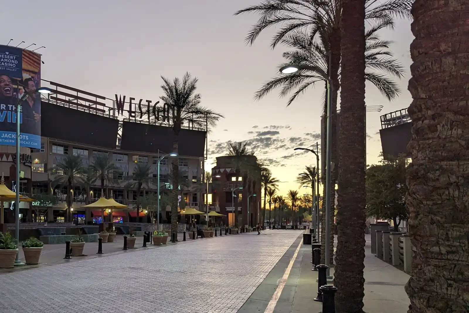 The Westgate district in Glendale, Arizona at sunrise, palm trees in silhouette along an empty brick paseo beneath the Westgate sign